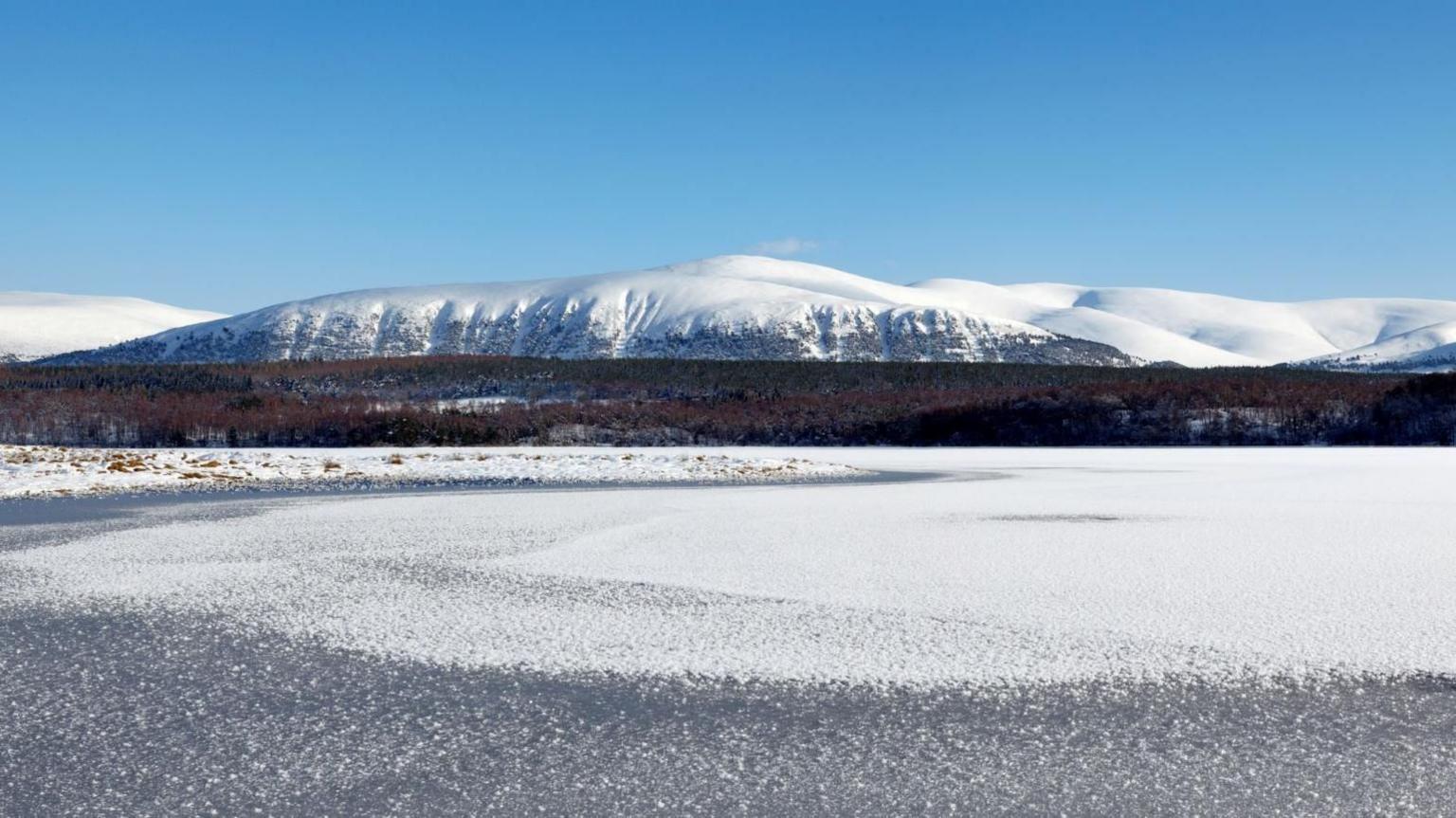 An area of water is frozen over and it is forestry and snow-covered mountains.