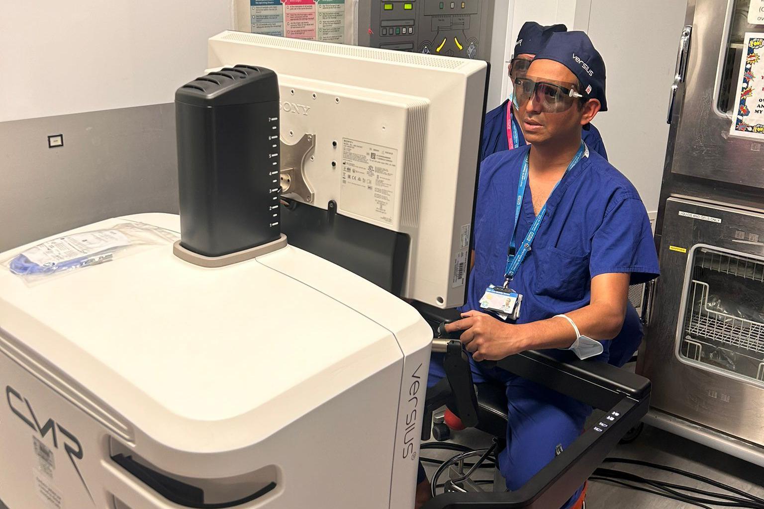 Consultant surgeon Pankaj Mishra is seated at the open console of the Versius Surgical Robotic System, wearing surgical scrubs, a cap, and protective glasses.