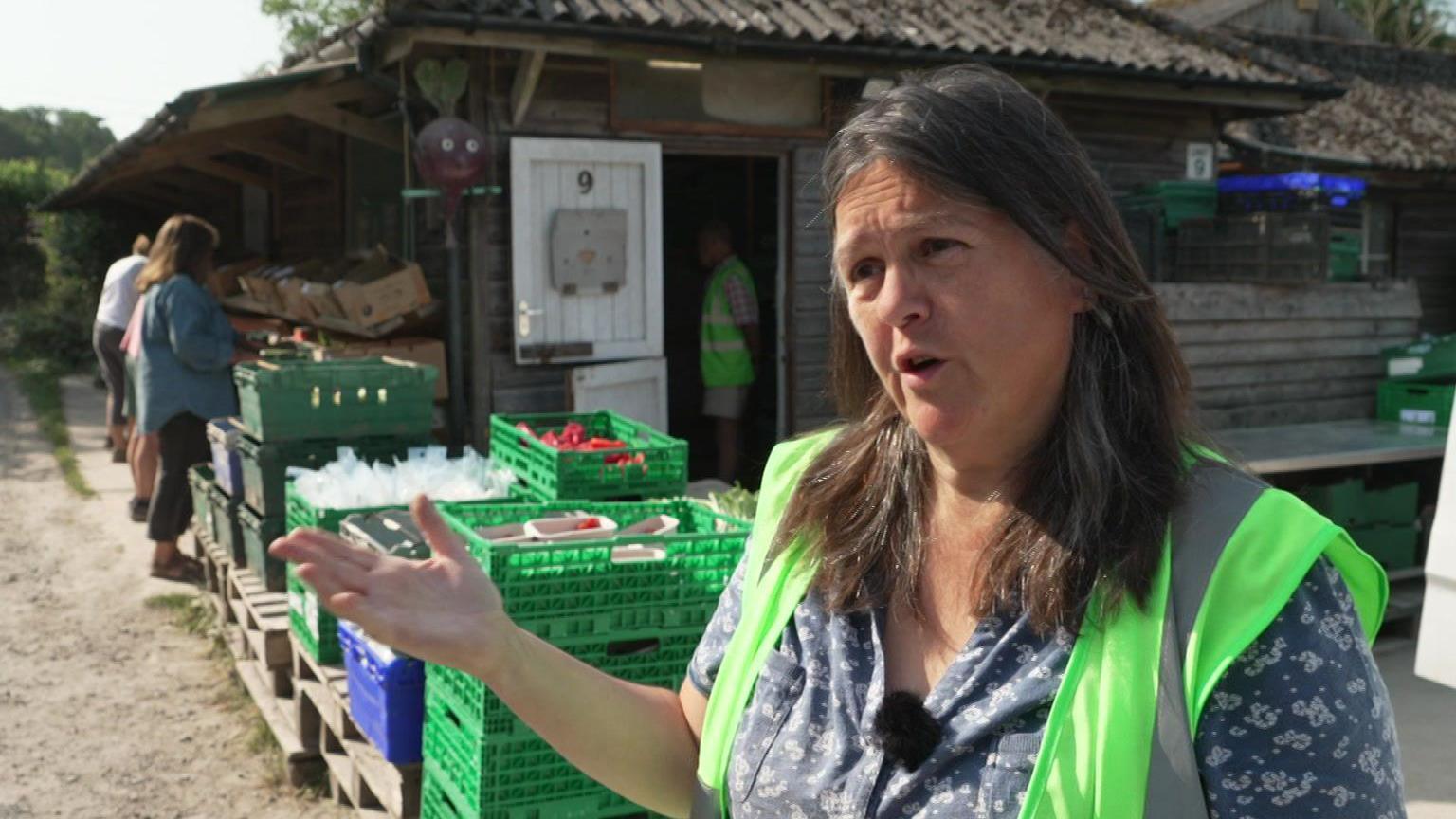 Sandra Crawford, the box manager for Food in Community, stands in front of rows of crates filled with fruit and vegetables. Behind her, volunteers are busy packing fresh produce into delivery boxes. She has shoulder length brown hair and is wearing a short sleeved blouse beneath a lime green high visibility vest.