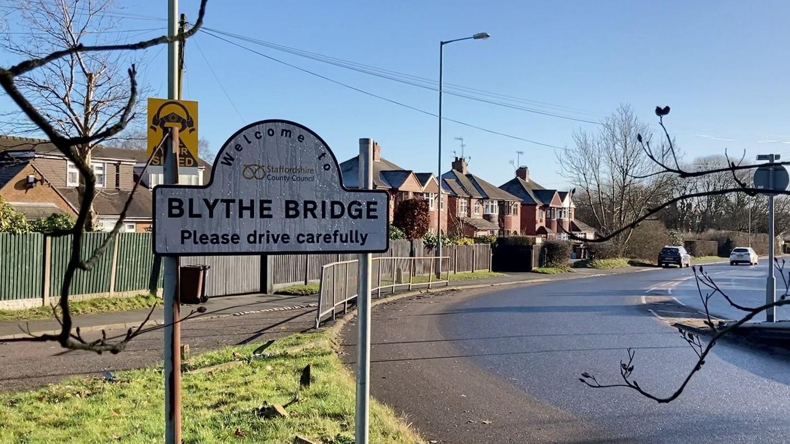 A sign on the left of a road reads: Welcome to Blythe Bridge, please drive carefully. A series of homes are in the background on the left