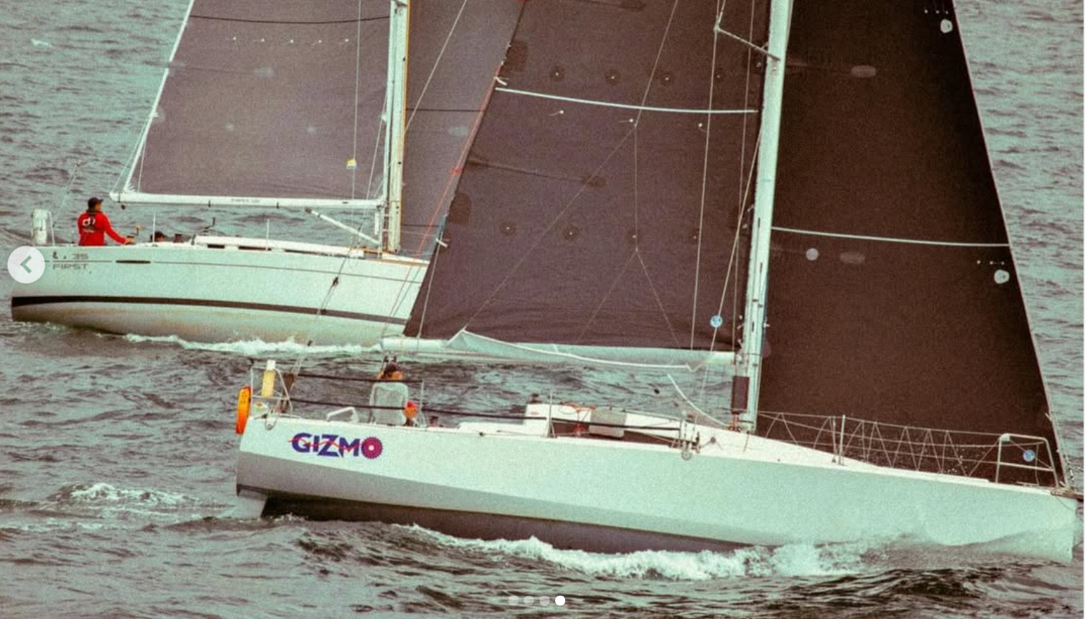 A yacht with the name Gizmo on the starboard side and a woman at the helm with dark sails and another yacht behind with a skipper in a red top.