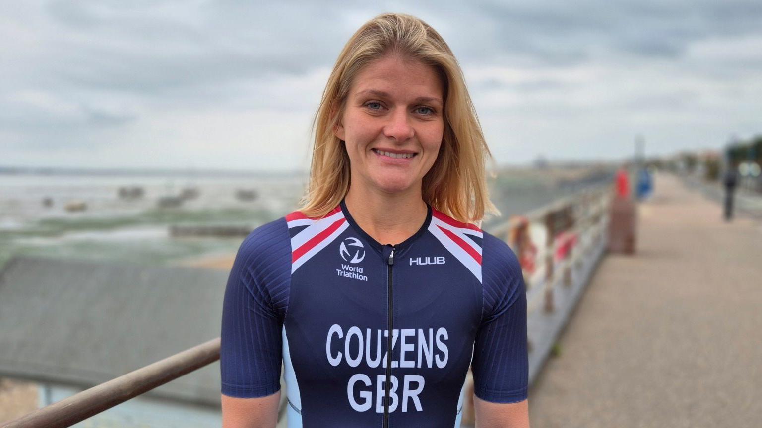 Georgia Couzens has shoulder-length straight blonde hair. She is wearing a navy blue skin-tight top with her surname and the letters GBR written in bold white font on the front, as well as a logo for the World Triathlon Championships. There are red and white detailing to her shoulders. Georgia is stood on Southend seafront with beach huts in the background.