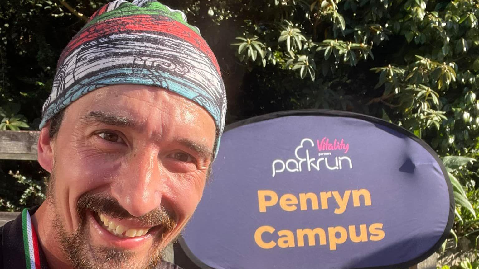 Adam Holland smiling and wearing a stripy bandana, stood in front of a purple Penryn Campus Parkrun sign.