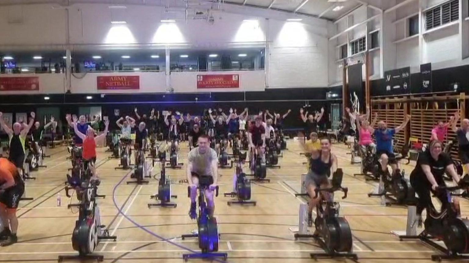Rows of static bikes in a large leisure centre hall, with the riders all waving