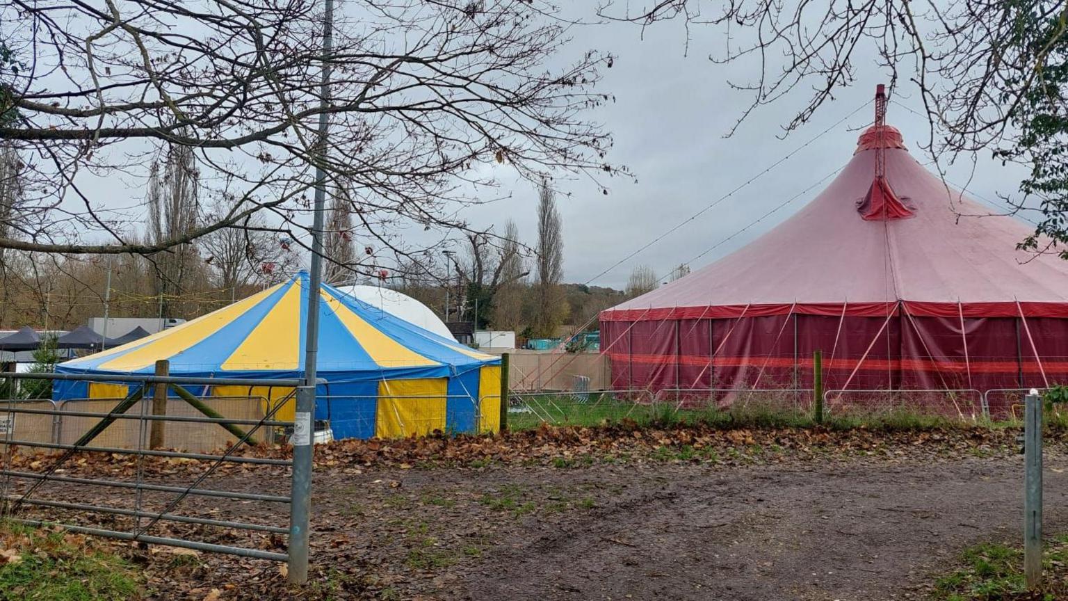 A gateway into a field where two circus-style tents have been erected. There is a large red tent on the right of the picture and a smaller blue-and-yellow tent on the left. It is a very autumnal scene. Fallen leaves lie on the ground and the skies are grey. No Christmas decorations are visible. 