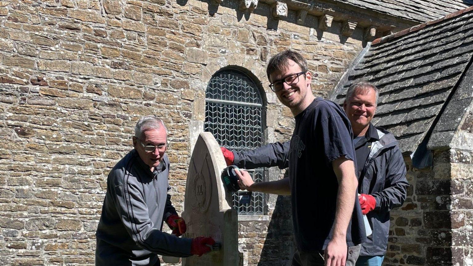 Prof Ed Coss, Dr Zack White and CSM Alexander McMillan turn to smile at the camera while each of them is working on different parts of the gravestone
