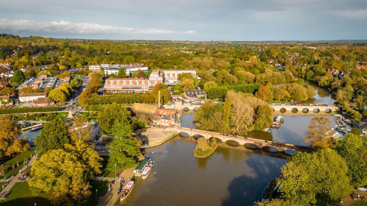 An aerial view of the centre of Stratford-on-Avon with trees on both sides of a wide river and large buildings seen between them. There are two large bridges crossing the river