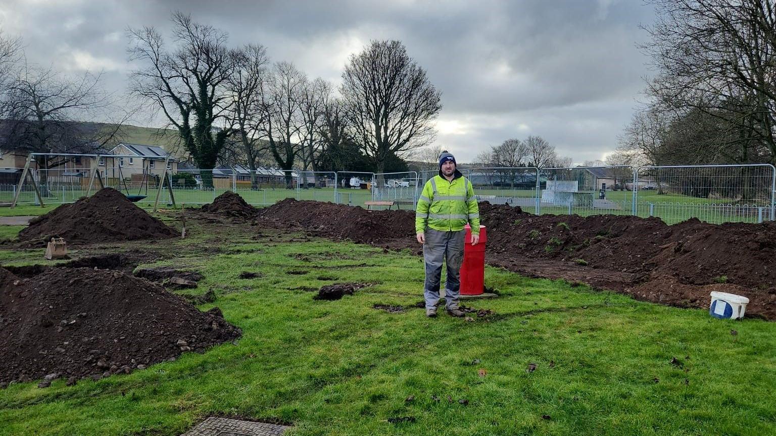 Mark Mather is standing in a park with lots of earth around him. He is wearing work clothes including a yellow hi-vis jacket and a grey woolly hat.