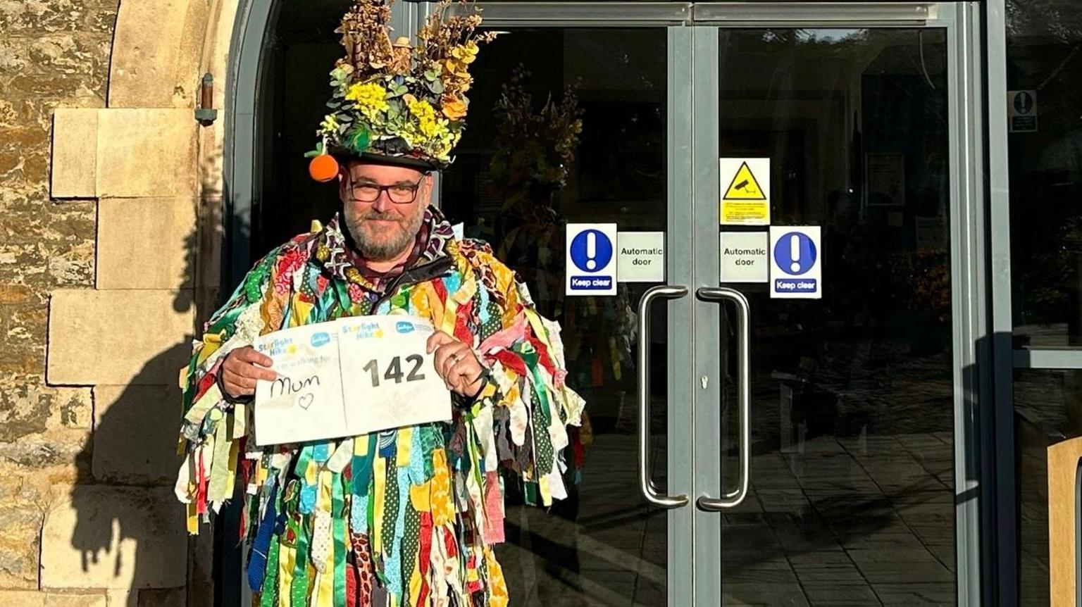 Peter, a man with glasses and a grey beard, wearing a tattered, colourful dress, holding a white letter which says for mum, smiling for camera.