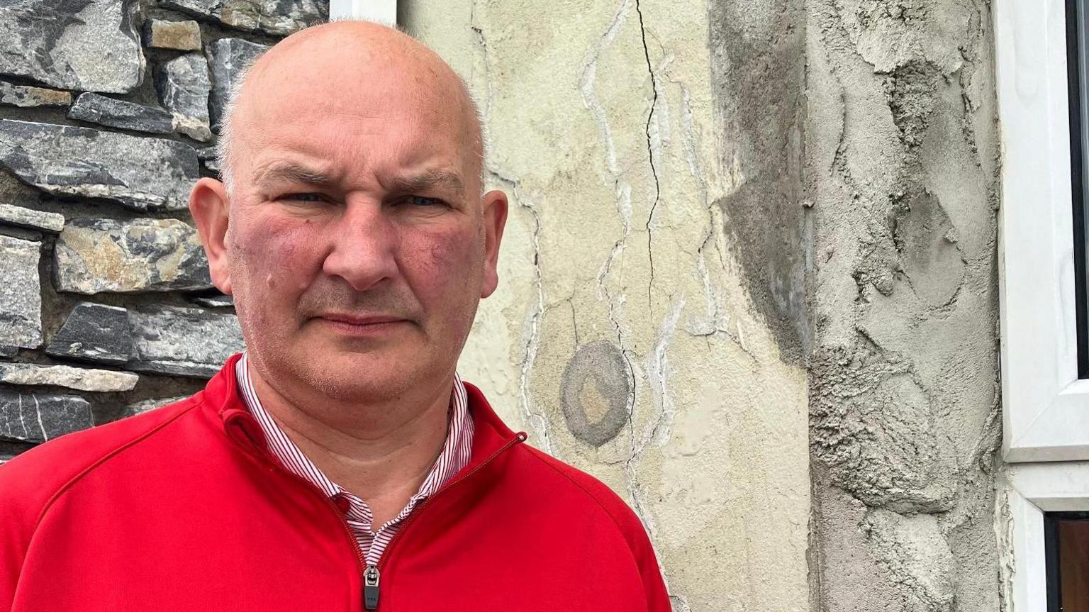 Ali Farren is wearing a red fleece and a white and red striped shirt. 
He is standing near a house with defective concrete blocks.