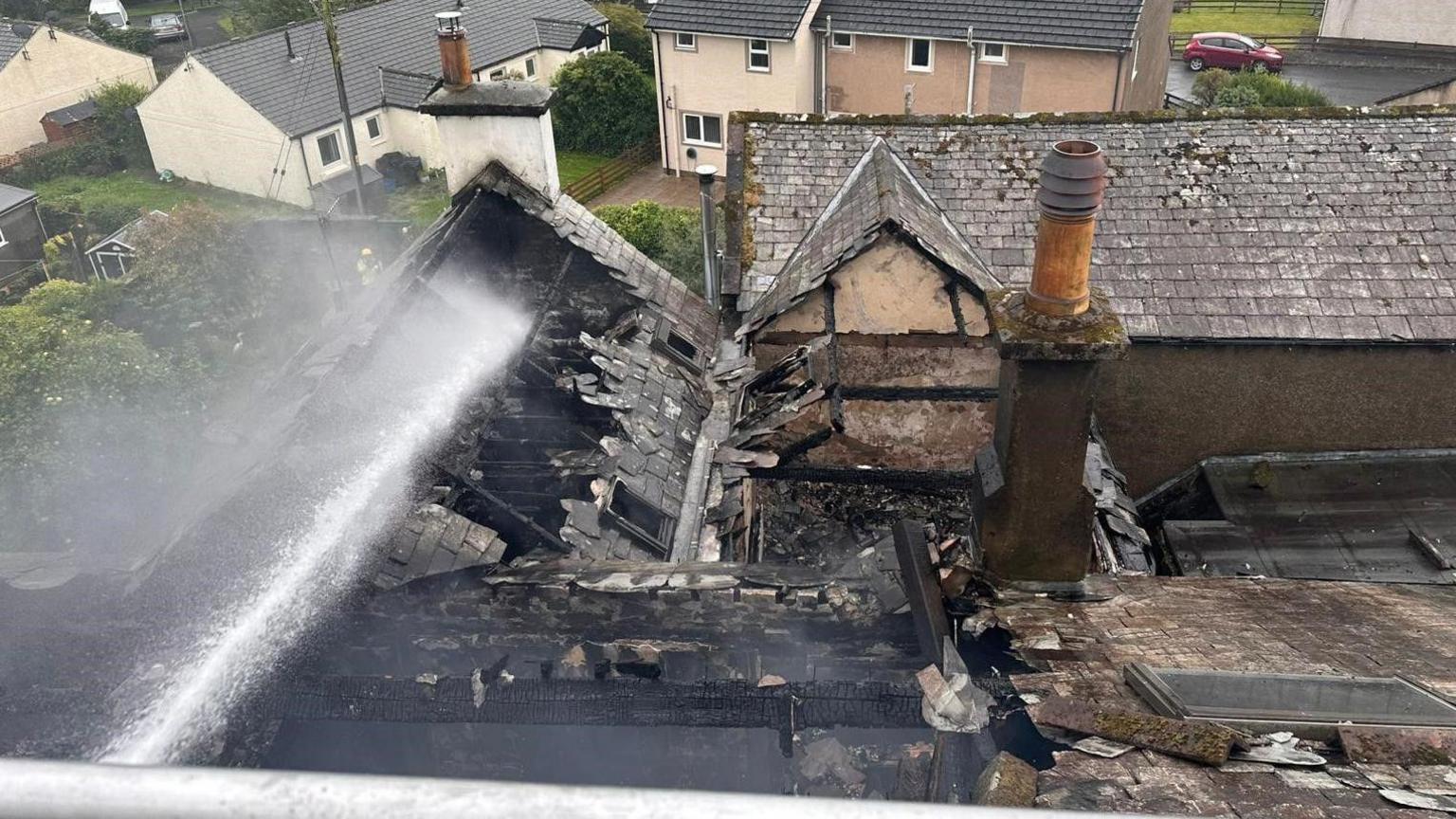 An aerial view of a building damaged by fire, showing a partially collapsed roof, charred timbers, and a firefighter hose jetting water.