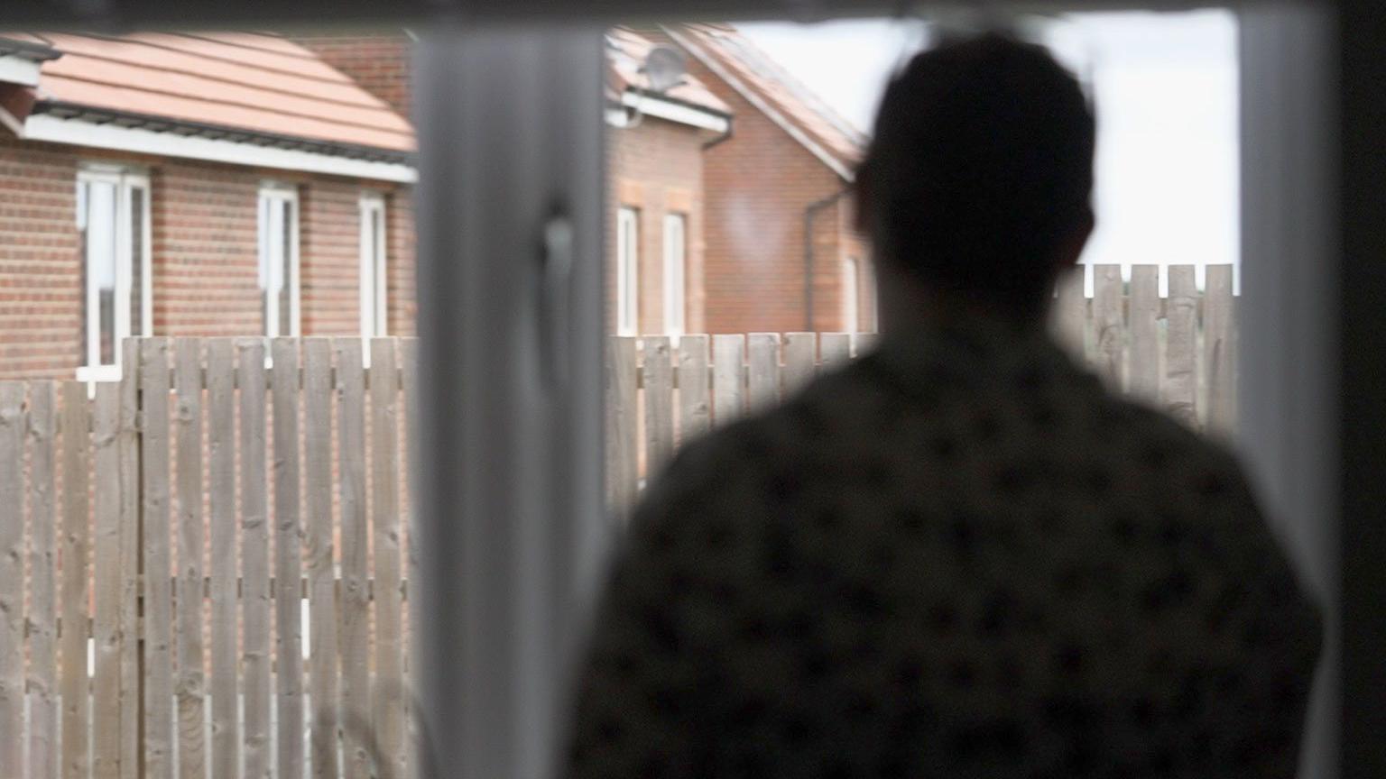 Anonymous image of a mother we are calling Caroline. She has dark hair. Her image is blurred to protect her identity. She is looking though a window into a back garden with a wooden fence and a row of newish houses in the background.