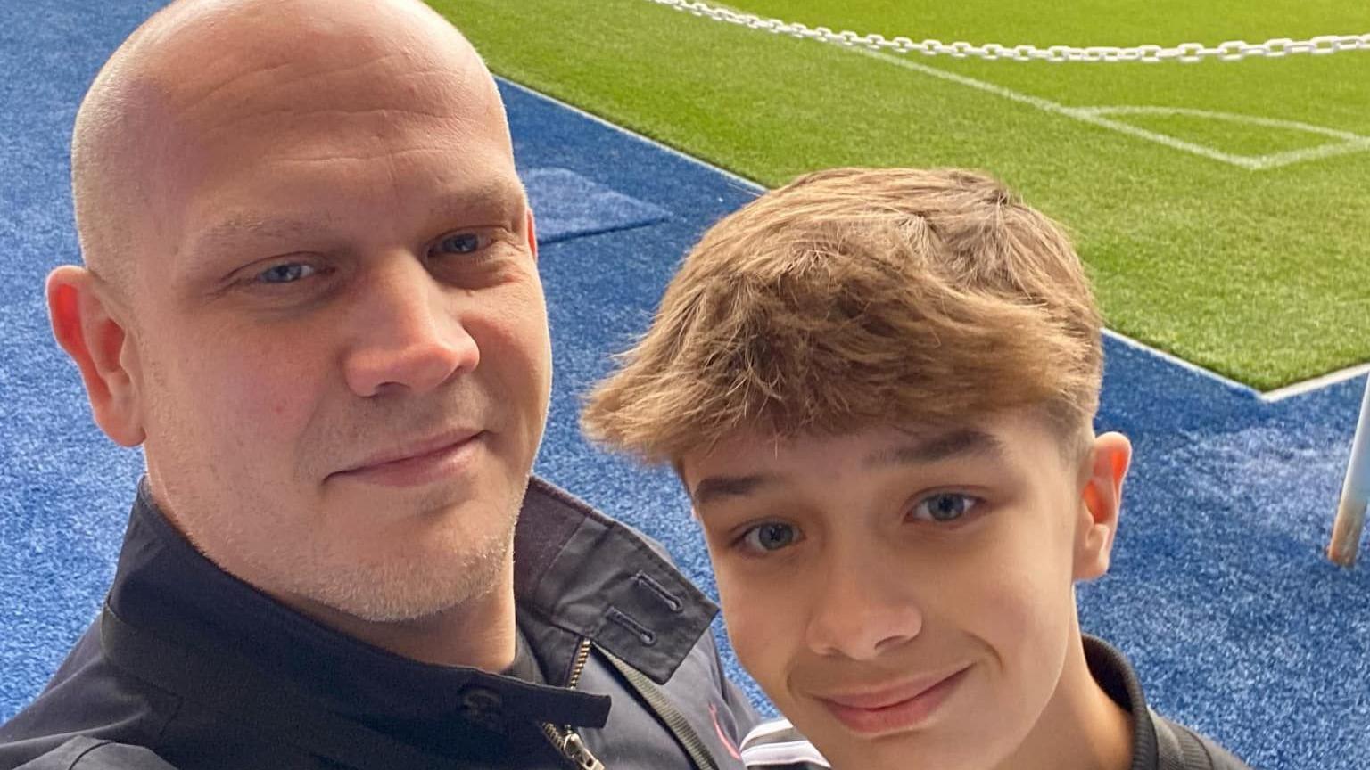 A bald man takes a selfie with a young boy stood next to him. They are smiling at the camera while standing at the edge of a football pitch.