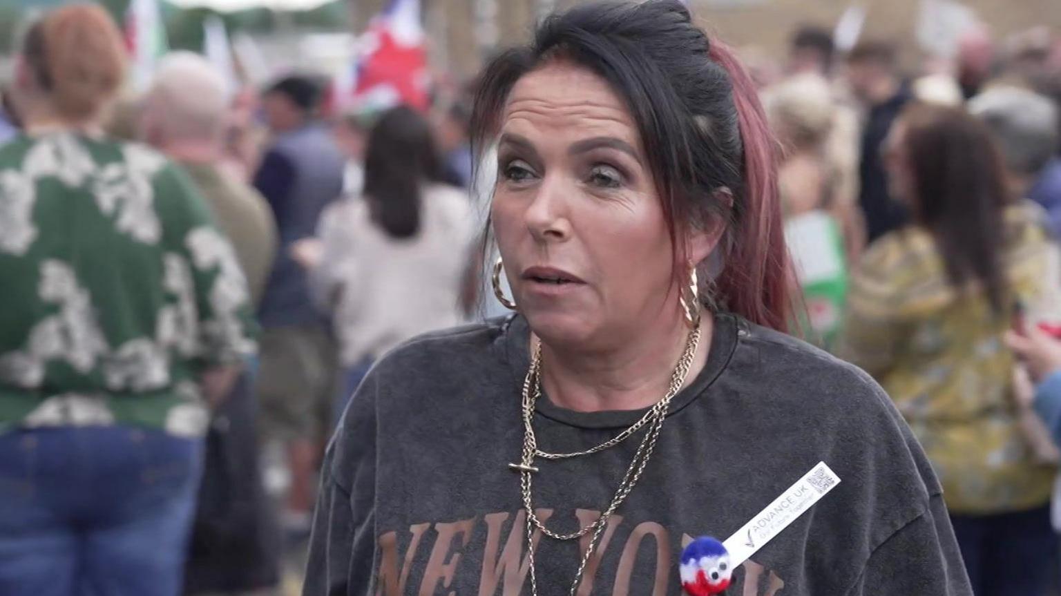 Netty Lloyd, 49, speaking to a journalist behind the camera. She had black and red hair, is wearing gold earrings and necklaces and is wearing a grey tshirt.