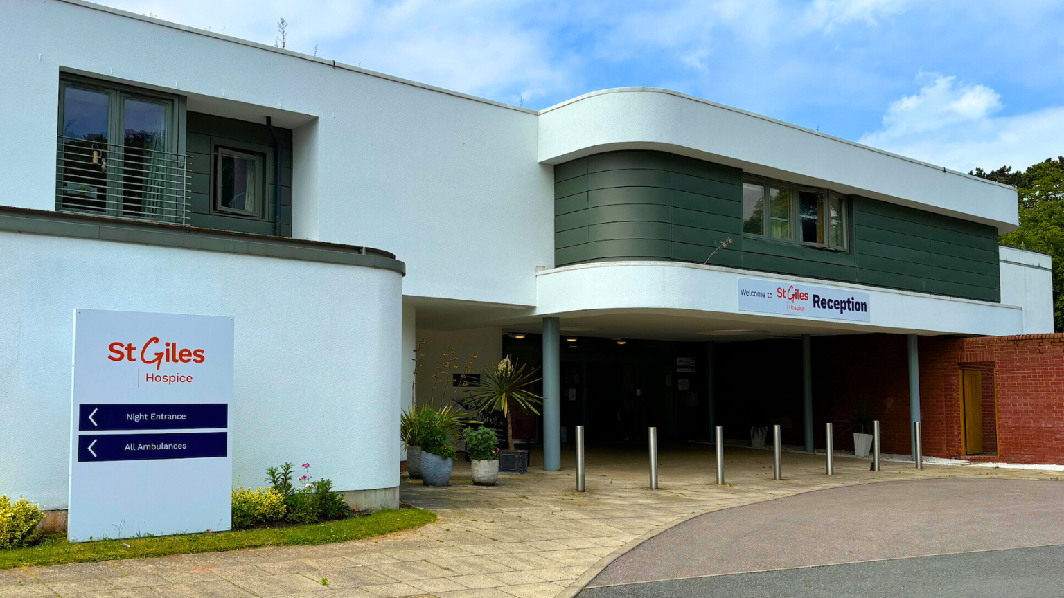 A white building with a sign to the left reading St Giles Hospice in orange. There are plants in front of the building and six metal bollards. 