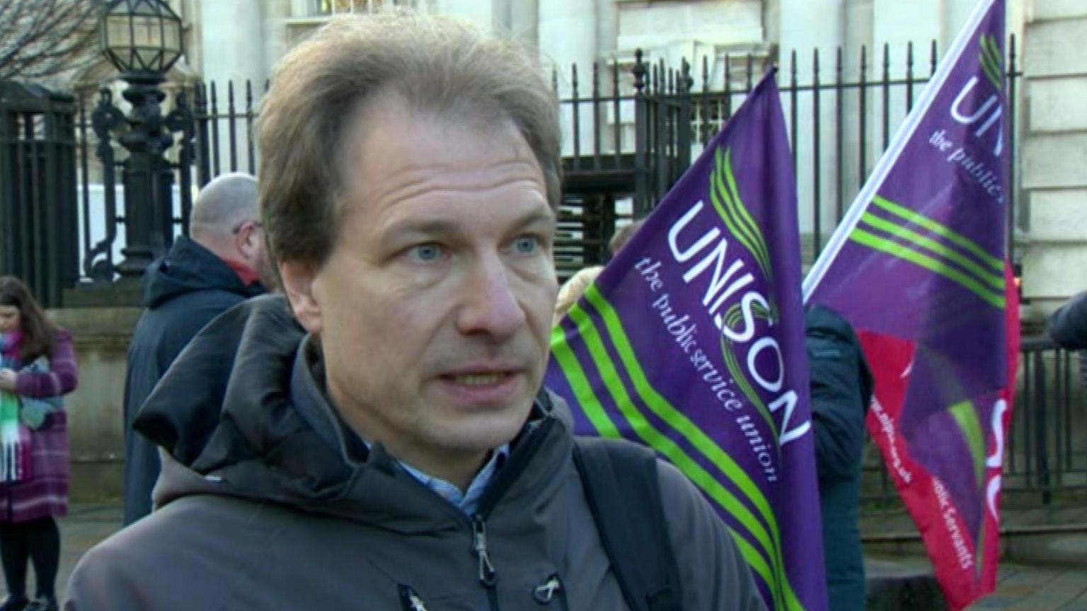 Daniel Holder has brown hair. He is speaking to the camera, wearing a grey jacket and backpack. There are purple Unison flags in the background. He is outside court. 