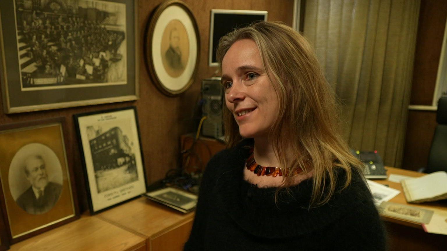 Emma Loat, who has long fair hair and wears a black top with a multicoloured necklace, speaks during an inteerview. She is in an office with black and white portraits of the previous family owners and venue displayed on the wall.