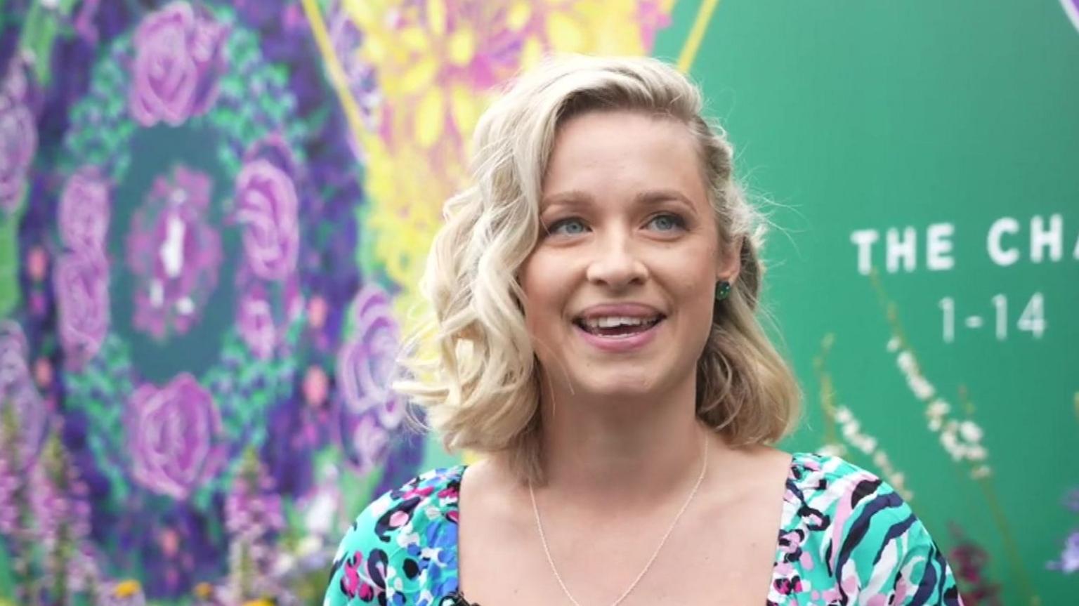 A head-and-shoulders image of a woman with short blond hair. Behind her is a mural, which is blue, yellow and green.