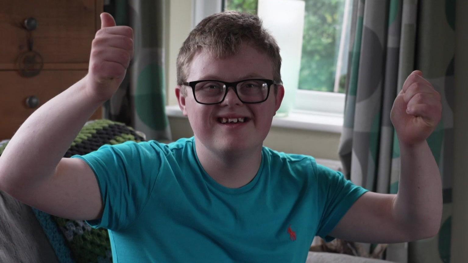 A teenage boy with Down's syndrome wearing a blue T shirt holds both thumbs in the air