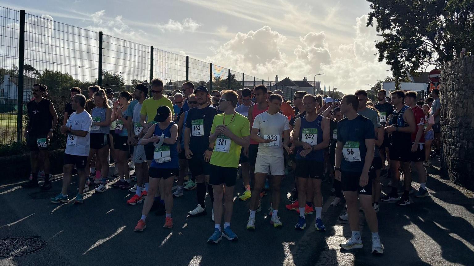 A group of runners ready to start the run. They are wearing running kit and have numbers pinned to the front of their tops. The sky has some blue sky and clouds but the runners are in shadow.