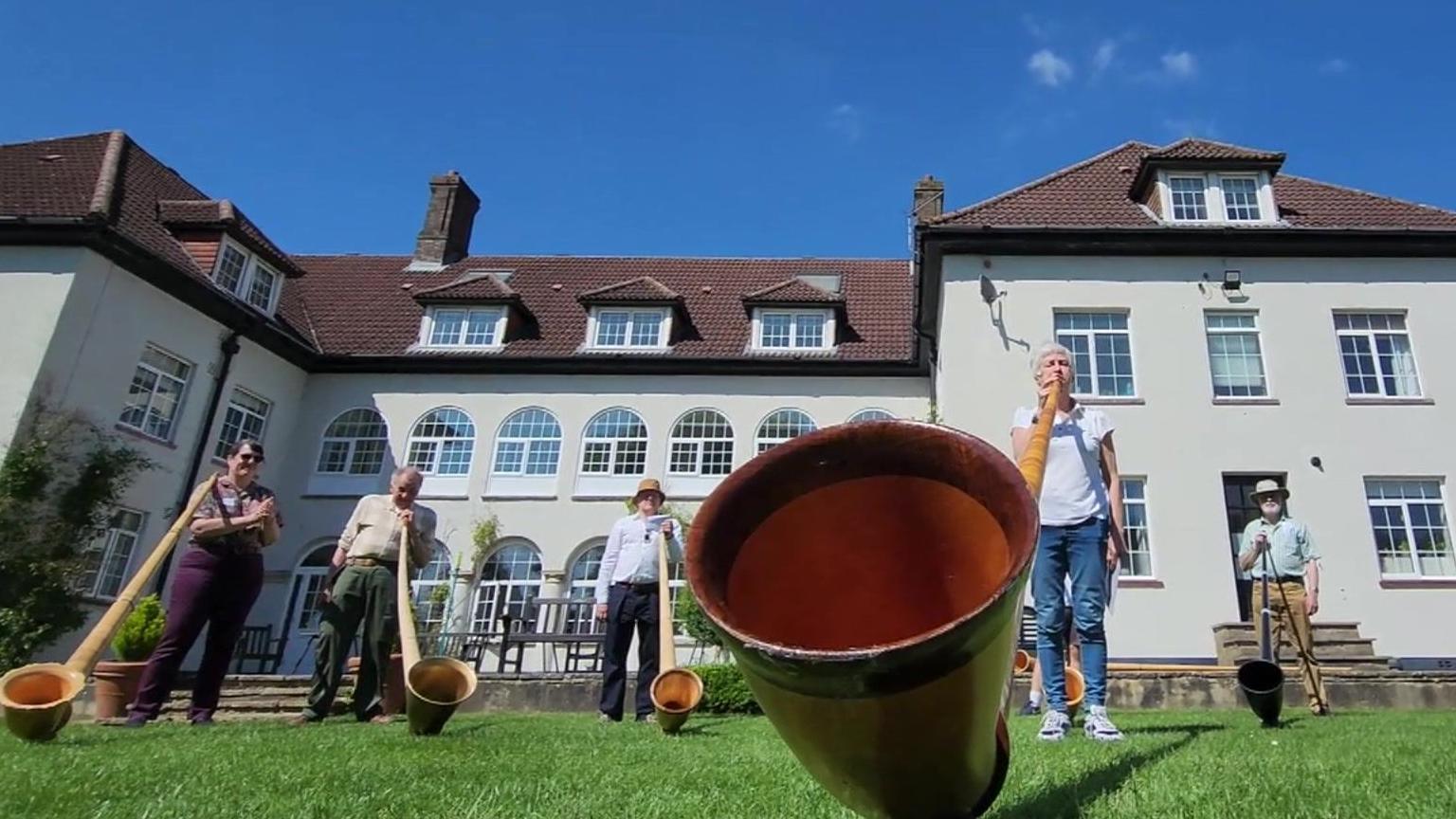 Row of five people playing alpine horns in front of a school building