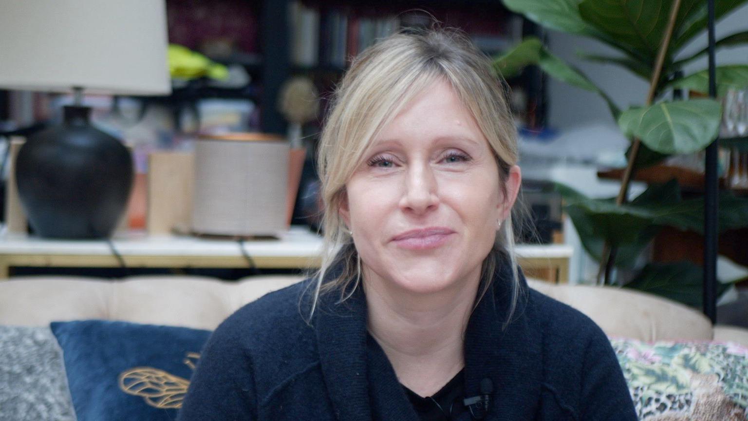 Dr Erin Sanders-McDonough is sitting on a sofa with brightly coloured pattern cushions. She is wearing a dark coloured top. 