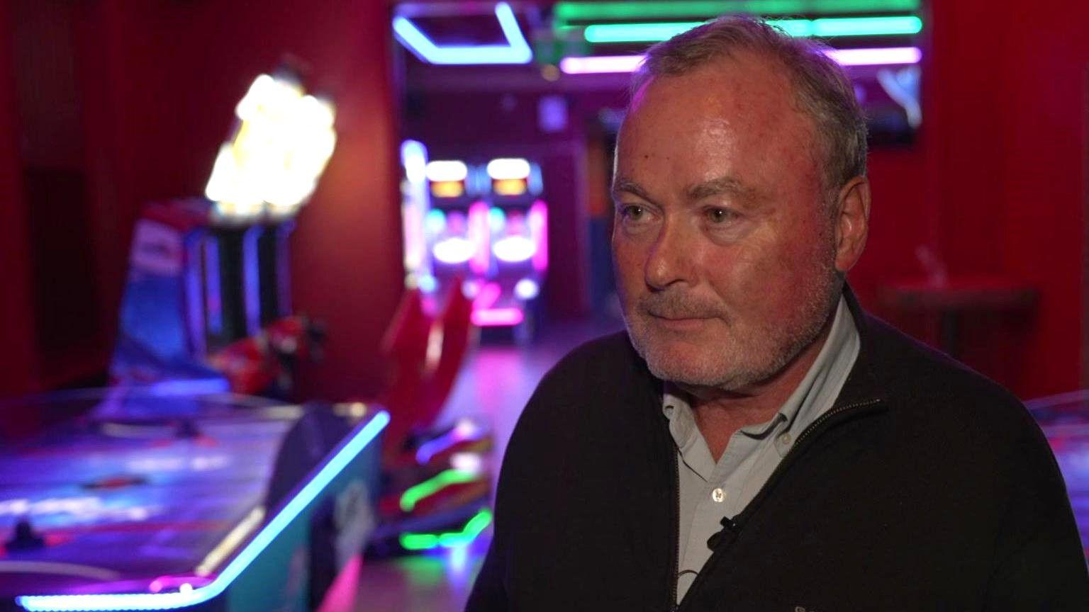 A man with short dark-grey hair and grey stubble stands in front of brightly lit arcade machines, which stand out in a club with low lighting. He is wearing a relaxed black fleece with a grey shirt underneath.