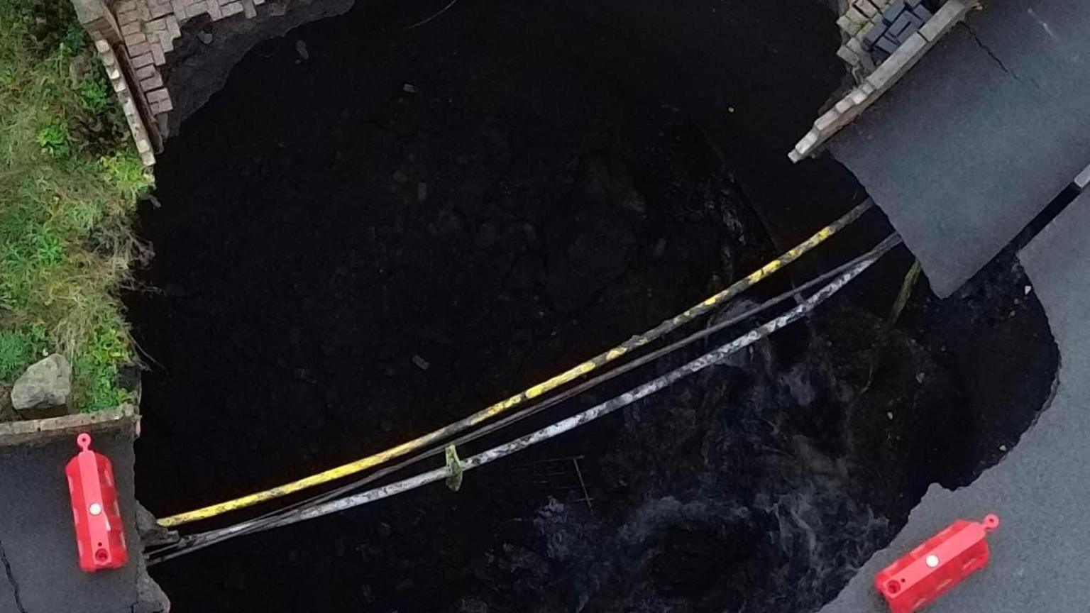 An aerial shot of the sinkhole in Nant Morlais, Pant, with pipes stretching across a large hole, and the remains of the road and pavement either side