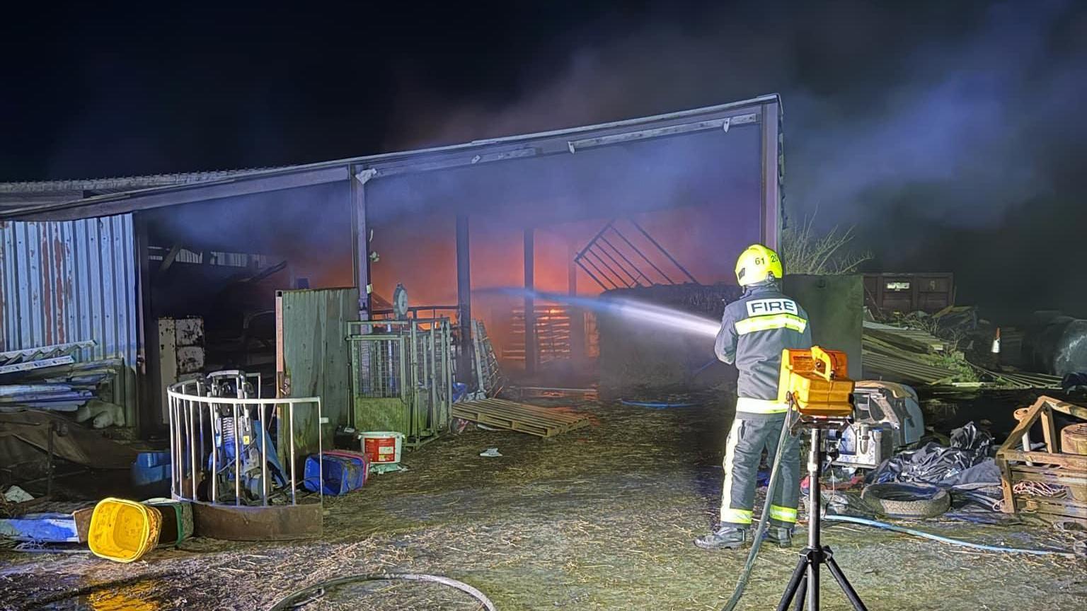 A firefighter is putting out a fire at a barn at night. The firefighter is wearing a black and yellow uniform and a helmet. The barn is burning with orange flames and thick smoke visible.