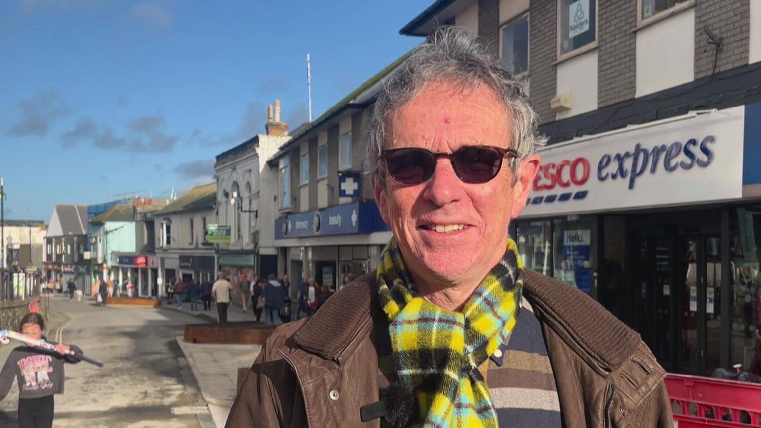 A man stands on the main shopping Street in Penzance - he's smiling in the sunshine - he wears a Cornish tartan scarf.