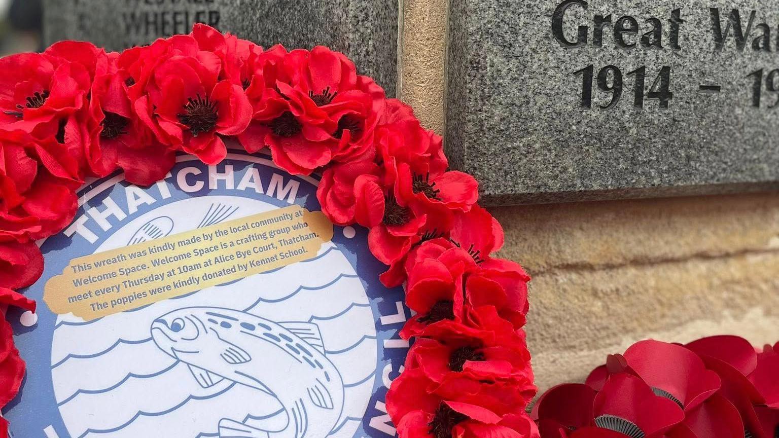 A close-up of a poppy wreath circling a fish image, laid at a war memorial. The words "Great War" can be seen carved into the stone.