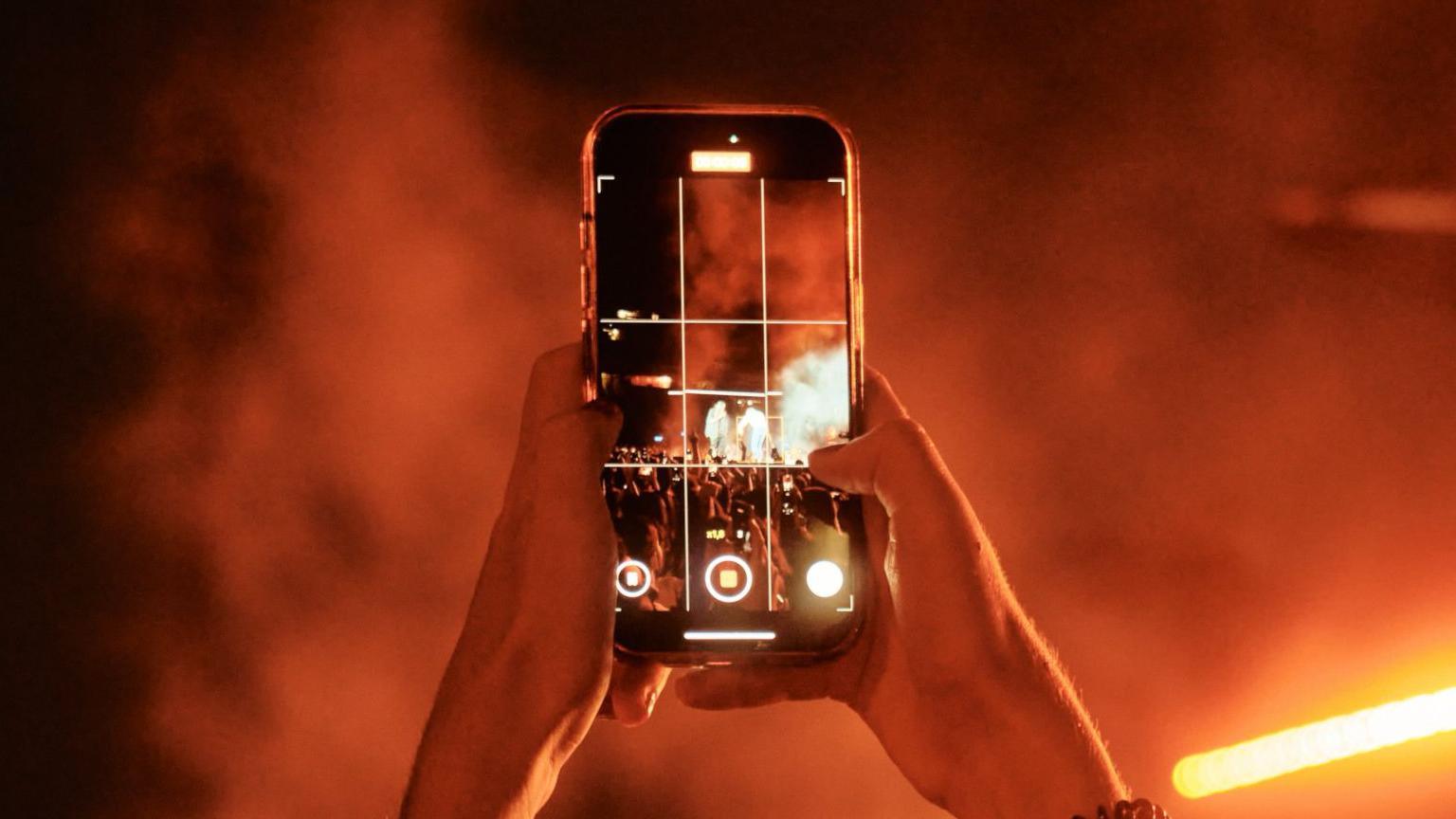 Somebody holding a phone up in a dark venue. Orange lights can be seen in the distance. The person is filming on their phone.