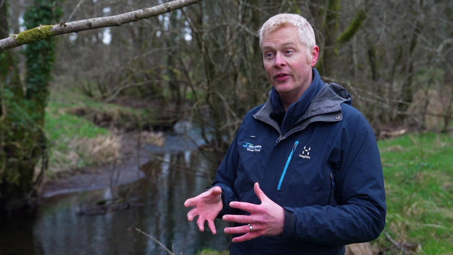 WCRT assistant director Luke Bryant stands in front of river talking to camera. He is wearing blue jacket. 