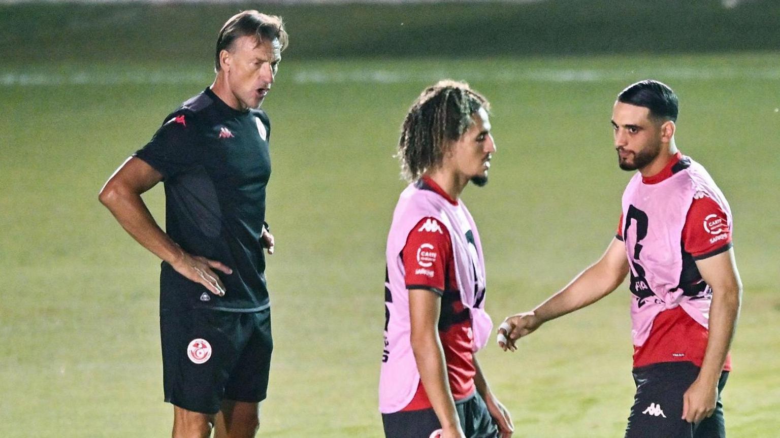 Herve Renard, wearing a black Tunisia training t-shirt and shorts, looks on as two Tunisia players in red shirts, pink bibs and black shorts take part in a training session on a green pitch