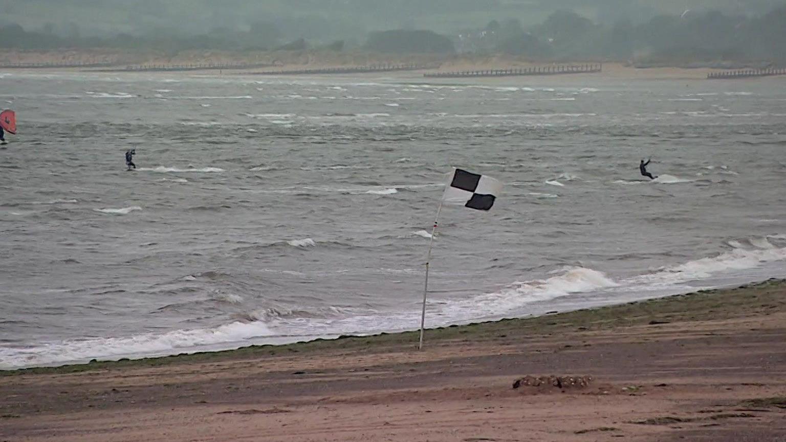 Two kitesurfers and a wingfoiler on the sea with a beach and a black and white flag.
