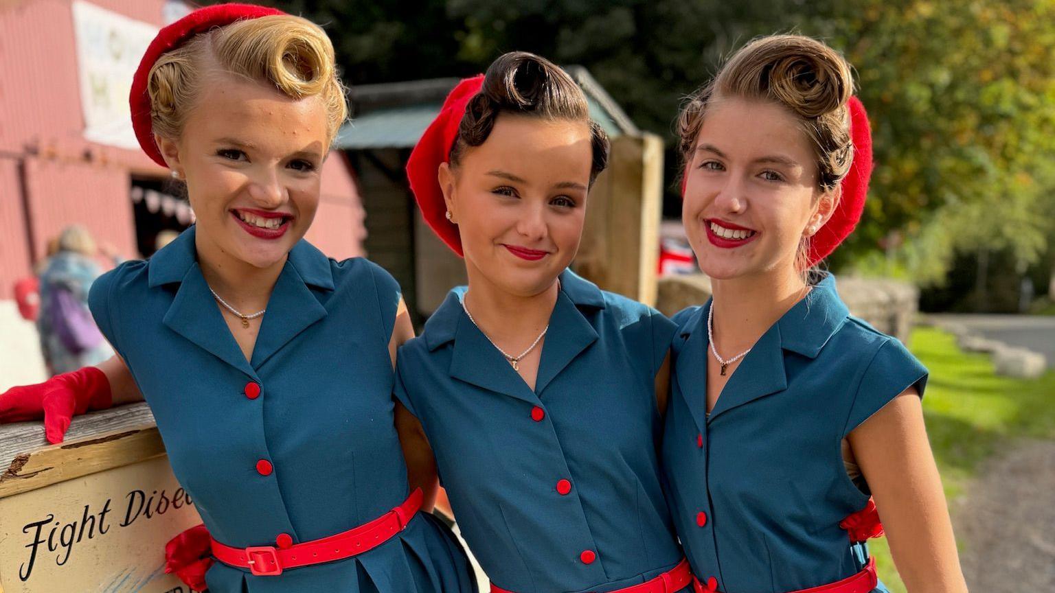 Siena, Emilia and Lottie smile at the camera. All three girls have long hair worn up with 1940s-style victory rolls and pink pill-box style hats. They wear blue 1940s tea dresses with bright pink buttons, pink belts and pink gloves, with red lipstick.
