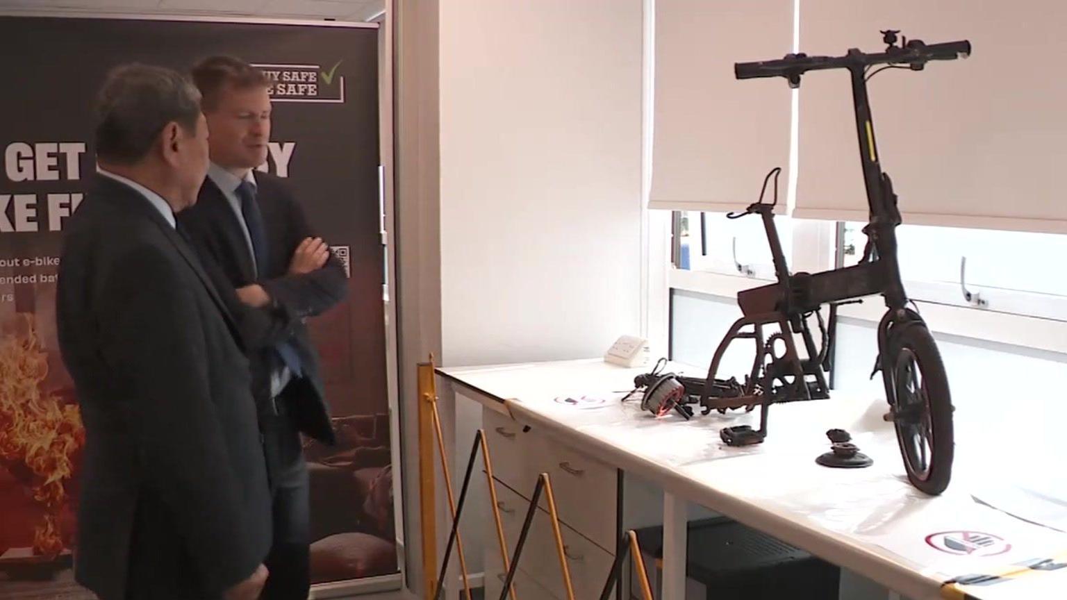 Product Safety Minister Justin Madders (right) stands and chats with another man while looking at a burnt out e-bike on display in an office