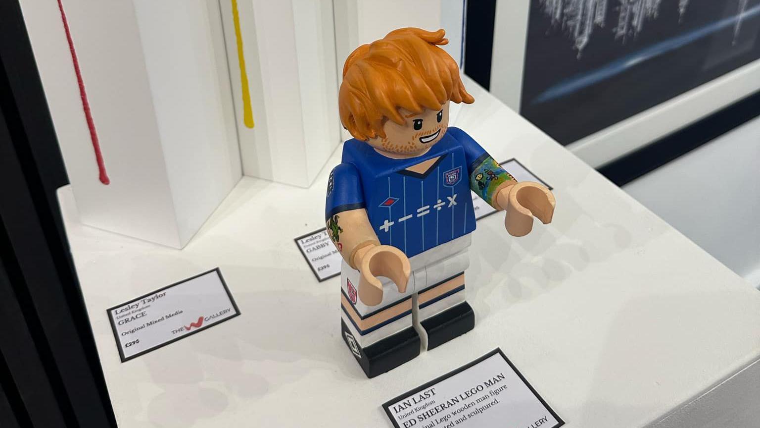 A view of the Ed Sheeran Lego figure from above. The whole figure can be seen. It is sitting on a white display table with an information card in front of it detailing information about the artist and creation. 