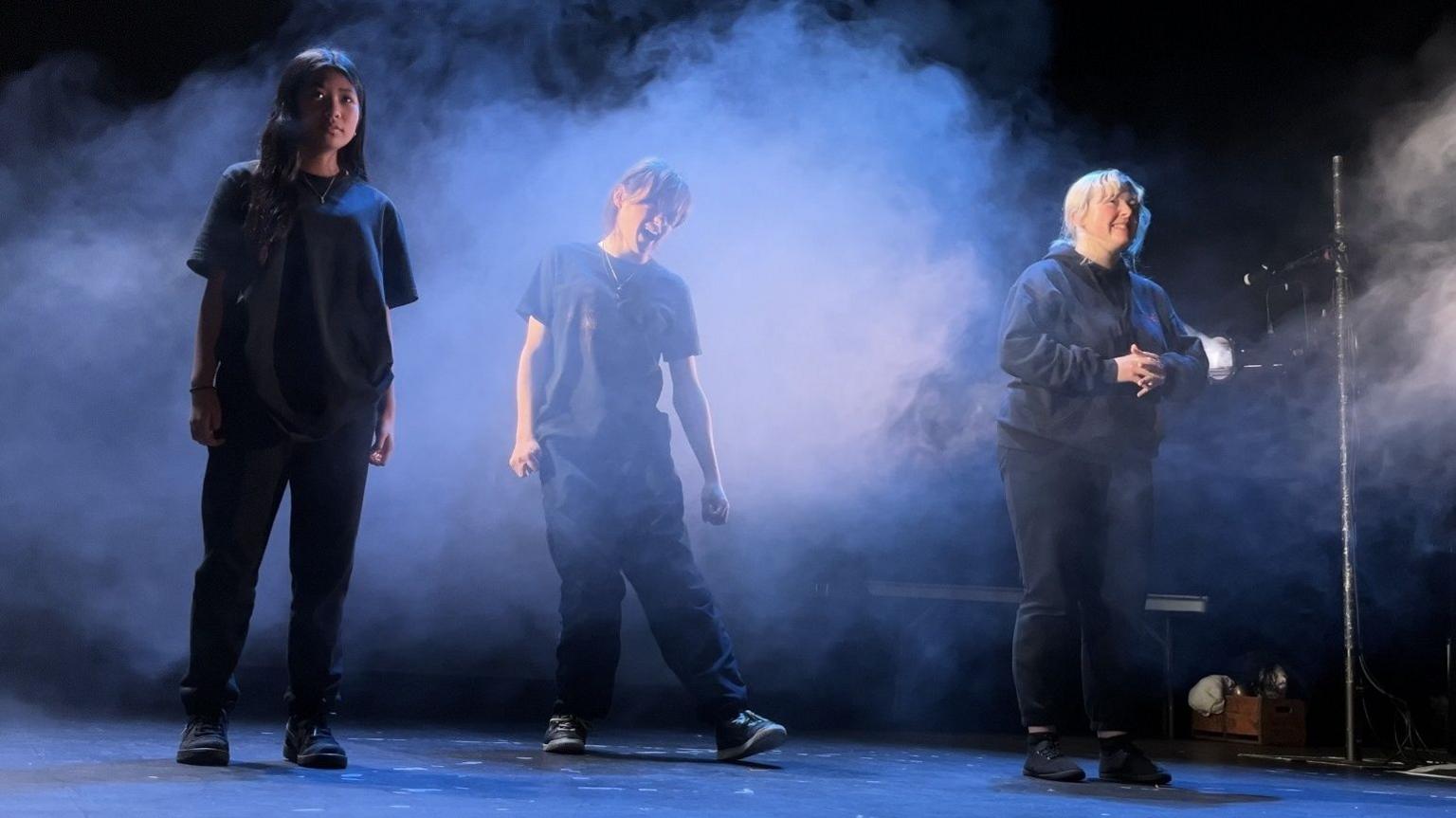 Three young actors from Prime Theatre dressed in black, dimly lit and surrounded by stage smoke