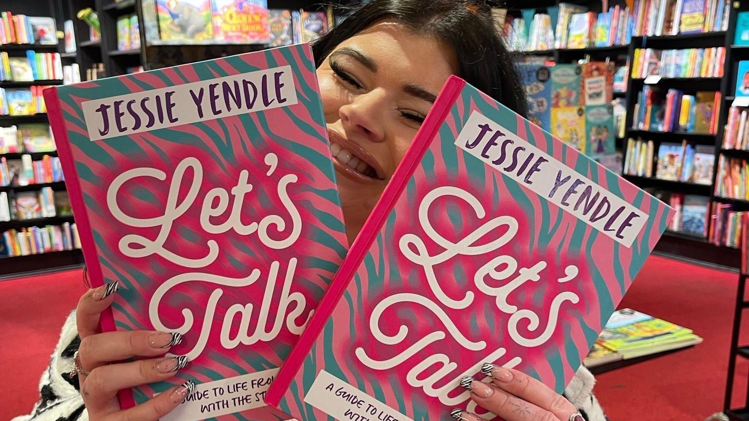Jessie seen smiling and holding two copies of her book inside a book shop.