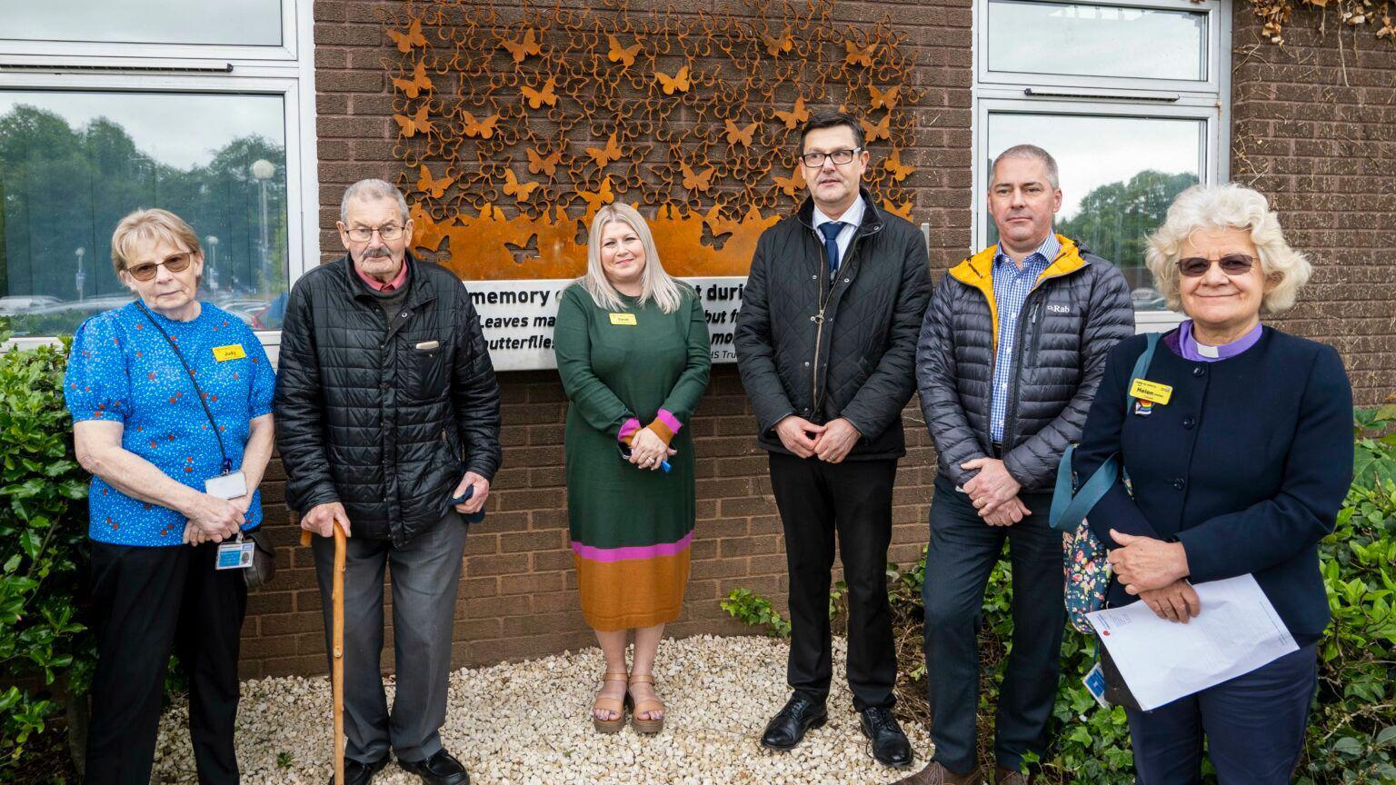 Staff standing in front of the steel memorial mounted on a wall, which has butterflies and a quote