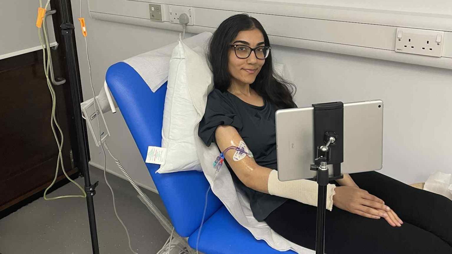 Sophie Akhtar, who has long dark hair and is wearing glasses, is sitting on a hospital trolley bed hooked up to a drip