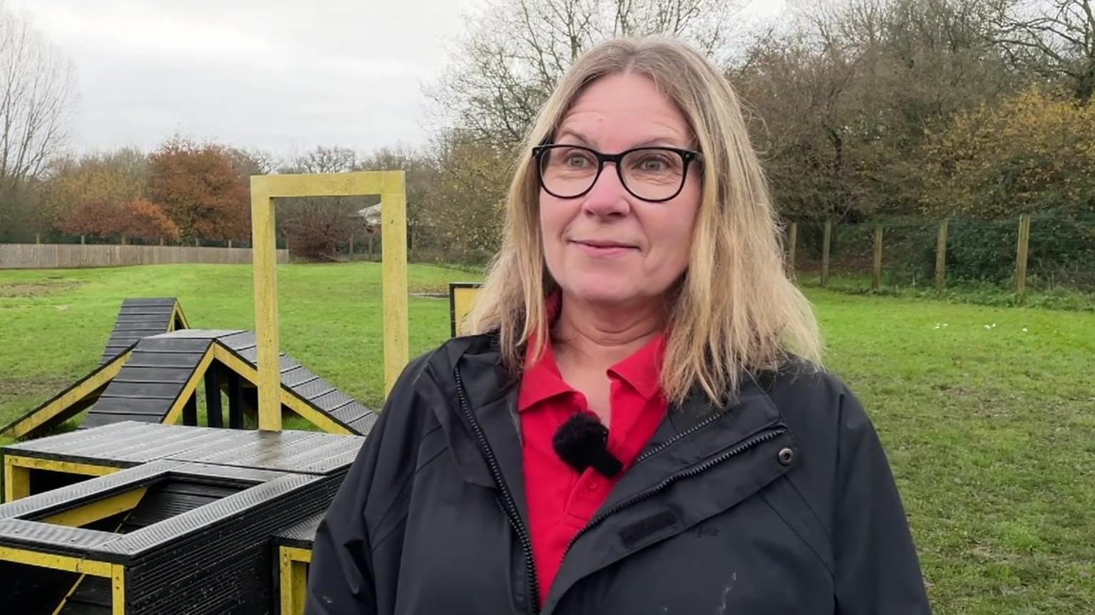 Diane is standing in a dog agility field with ramps and agility equipment behind her. She is wearing a black raincoat with a red collard top underneath and she has blacked rimmed glasses on.