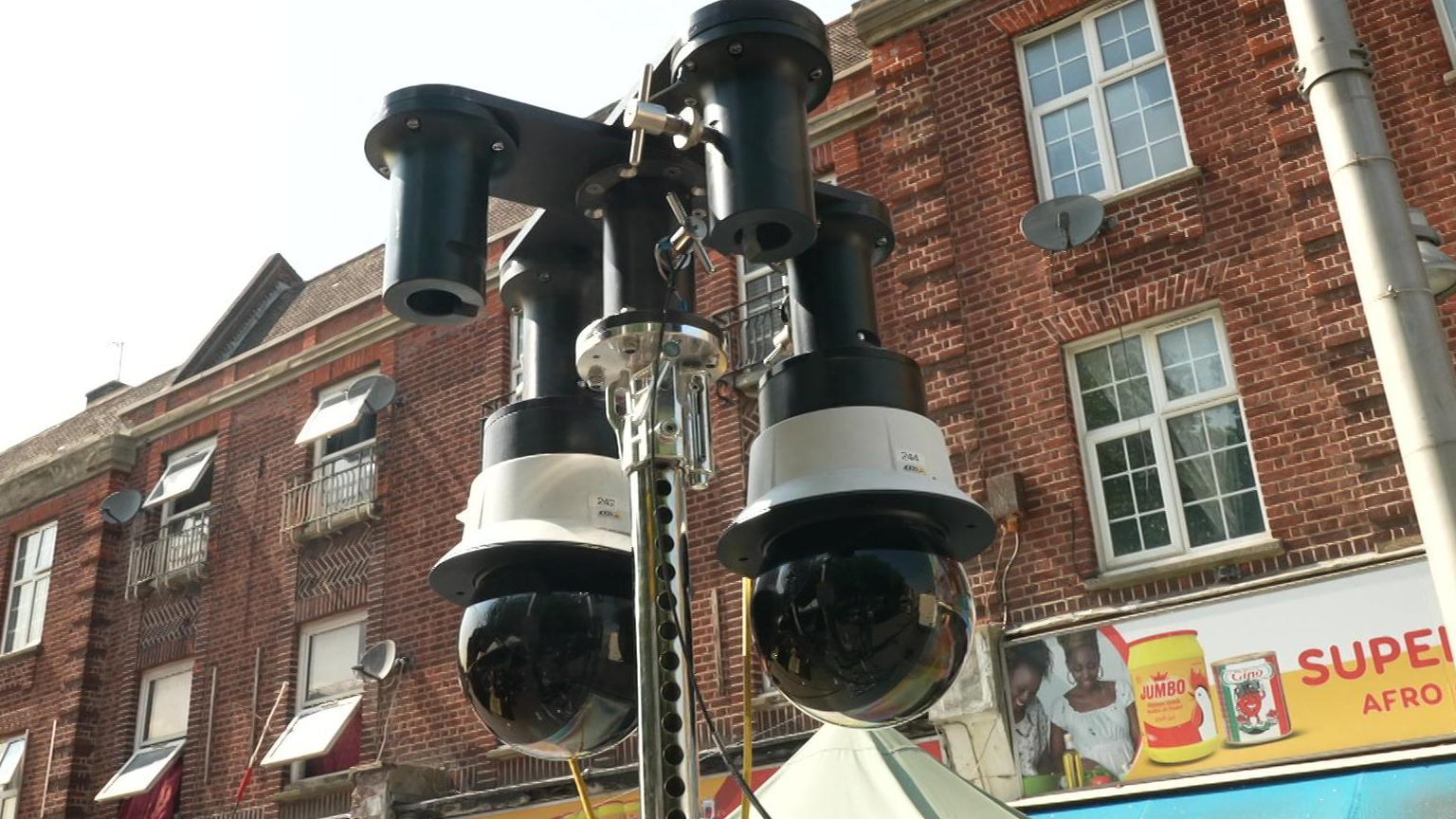 Two cameras, which resemble black globes, mounted on a pole, in Walthamstow market, a brick building and part of a shop sign in the background