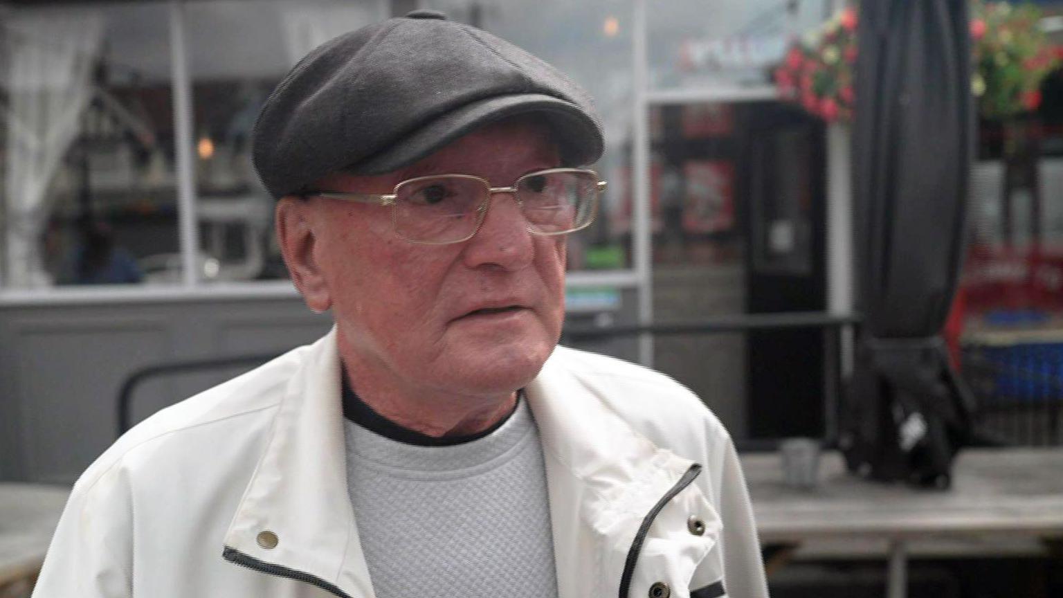 Paul is wearing a dark grey flat-cap on his head, rectangular metal glasses on his face, a white jacket over a grey top. He is standing in front of a grey pub building with floral hanging baskets.