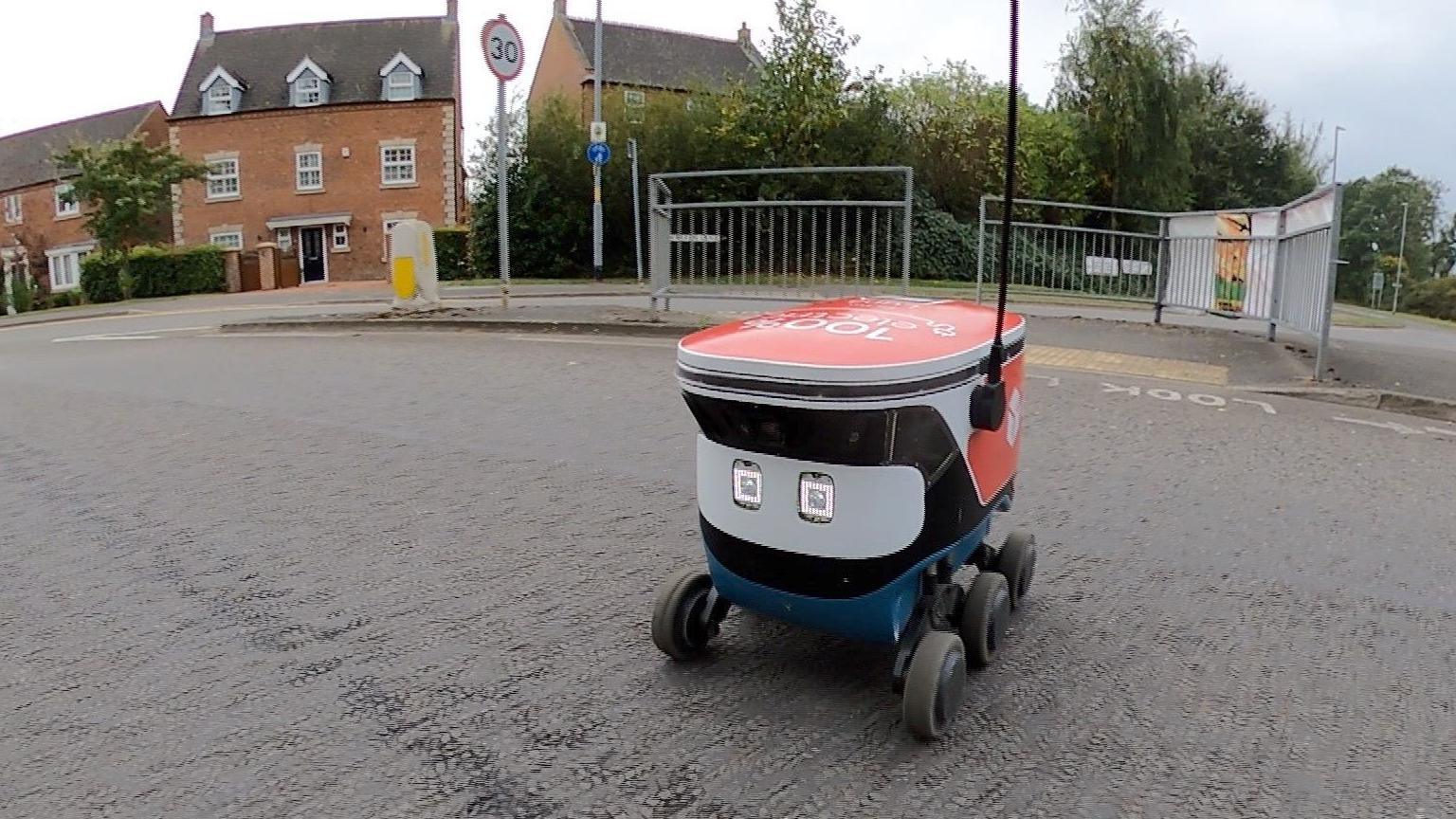 A small rectangular delivery robot with six wheels and headlights which resemble eyes drives itself across the road. The robot has a blue chassis and white body with red DPD branding, a black antenna is fixed to its left side and reaches into the sky.