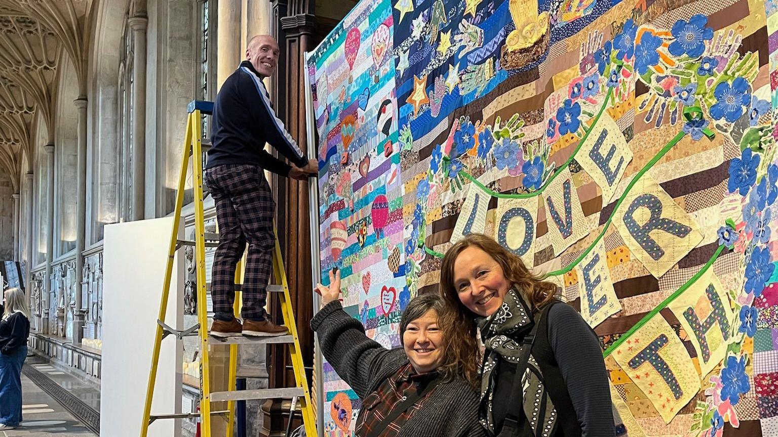 Jo and Kate stood next to each other in front of the panel, while a man nearby stands on a ladder to hang it