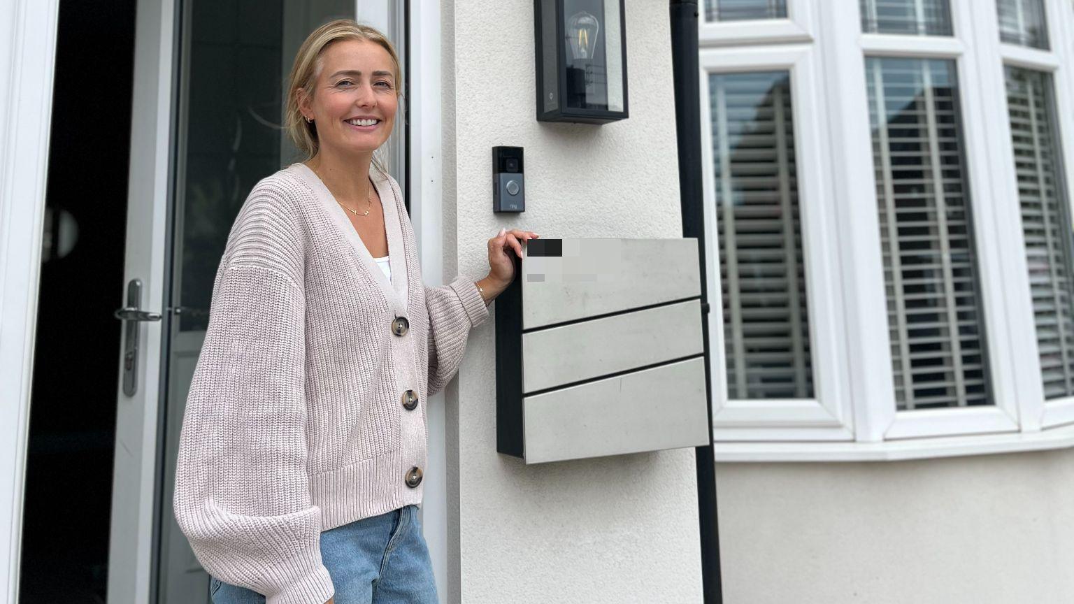 Rachel Roberts, who has blonde hair and a cream coloured hoody, stands on the doorstep of her semi-detached home which has white rendering.