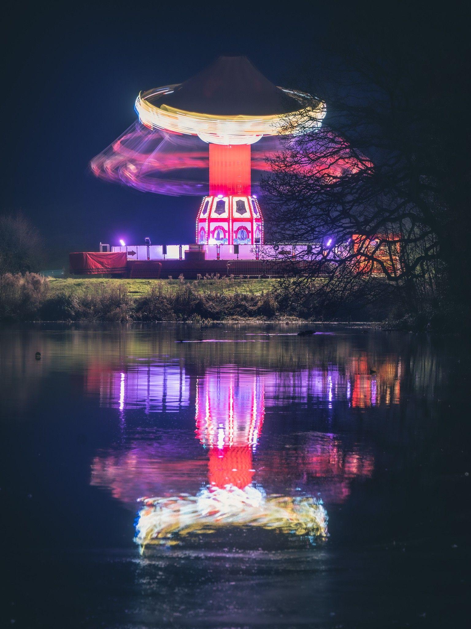 A whirring fairground ride is pictured against the night sky. It is reflected in a river which is in the foreground of the picture.