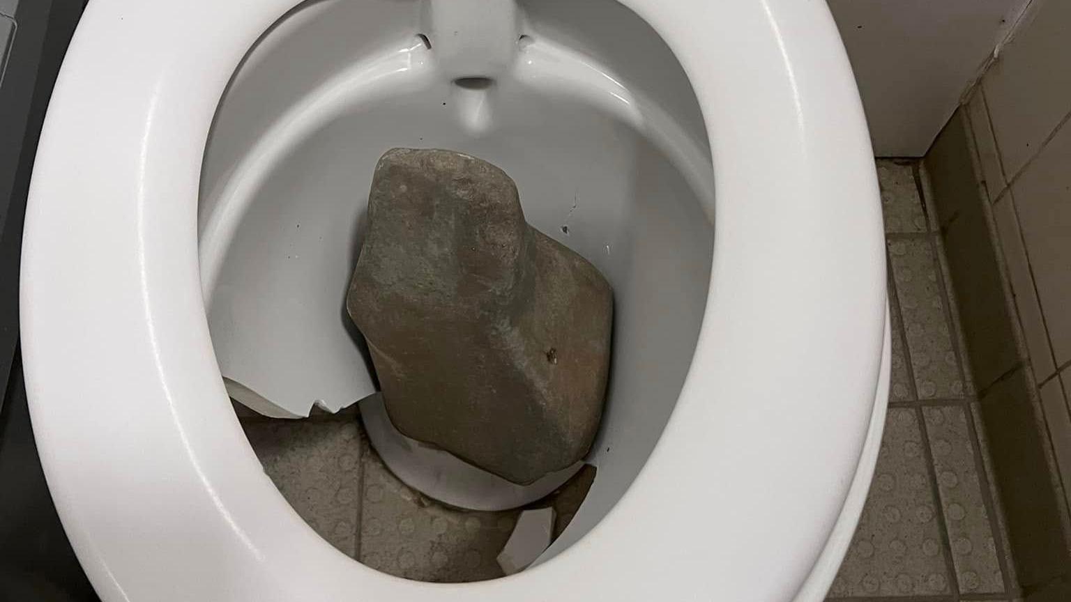 A broken toilet bowl with a large rock in it. The front of the bowl is completely broken and you can see the floor through it.